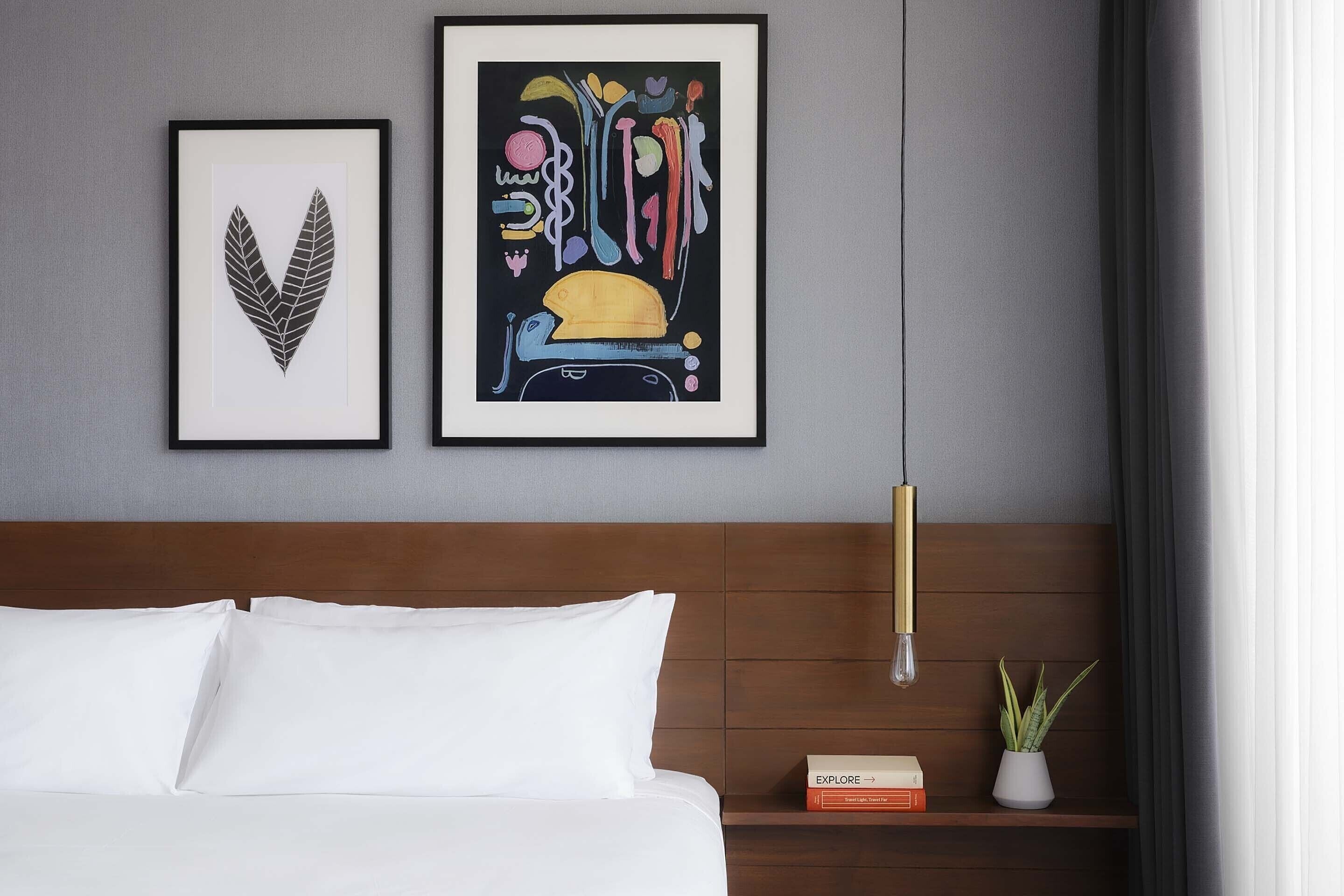 A neatly made bed with white linens, a wooden headboard, a framed abstract artwork, a small bookshelf with books, and a potted plant on a bedside table.