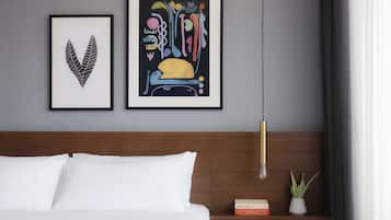 A neatly made bed with white linens, a wooden headboard, a framed abstract artwork, a small bookshelf with books, and a potted plant on a bedside table.