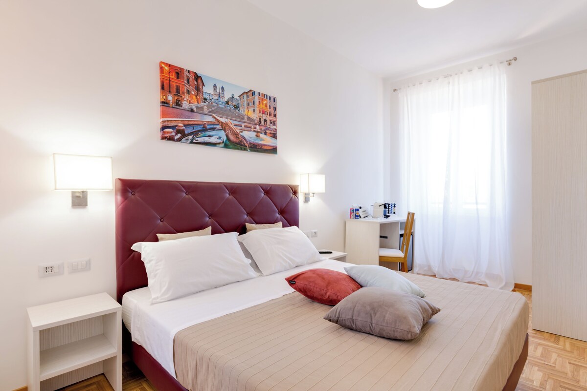 A neatly arranged bedroom with a large bed, a red headboard, white bedding, a nightstand, a bedside table, and a wall-mounted lamp.