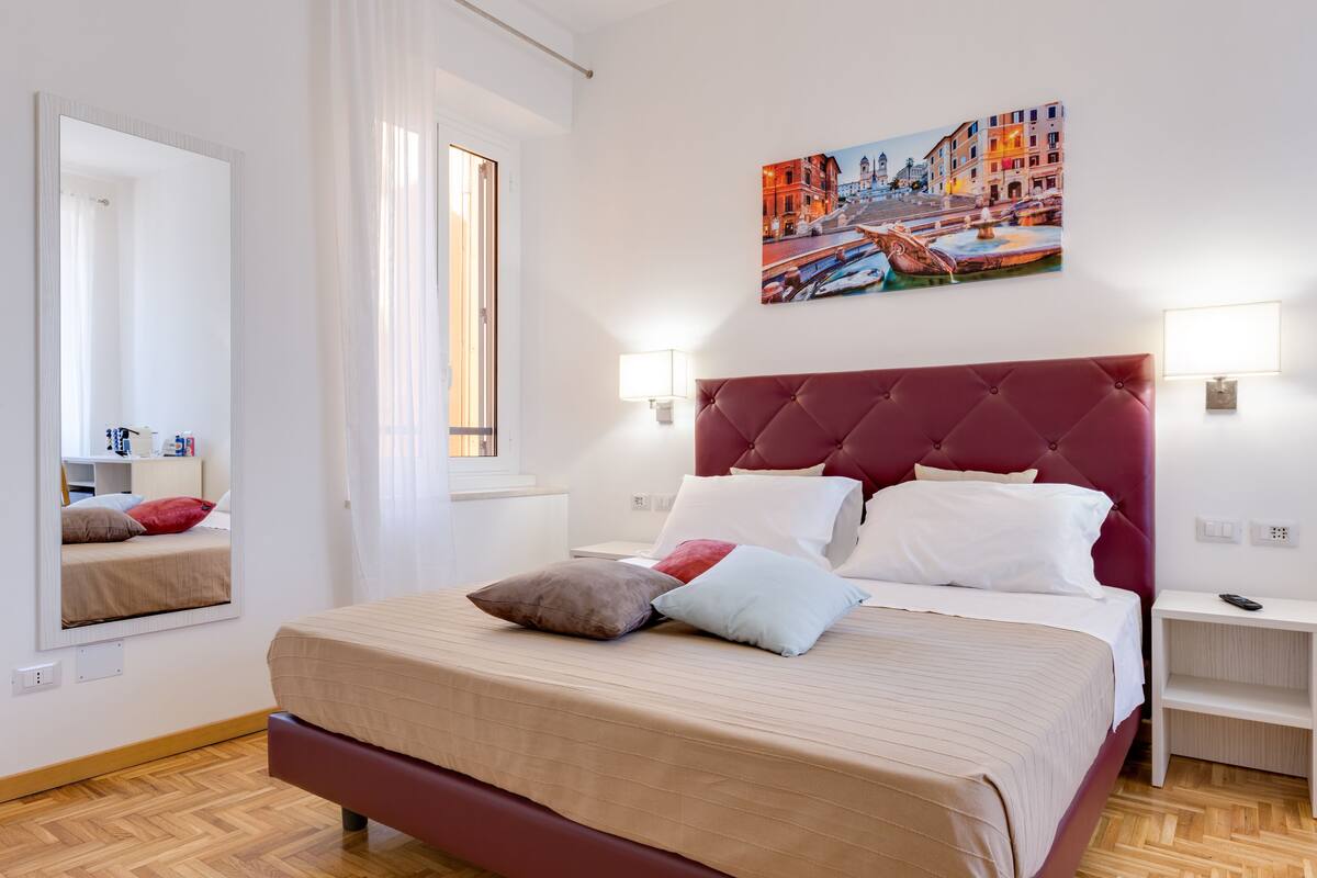 A hotel room with a large bed, a red headboard, a painting of a cityscape, and a window with curtains.
