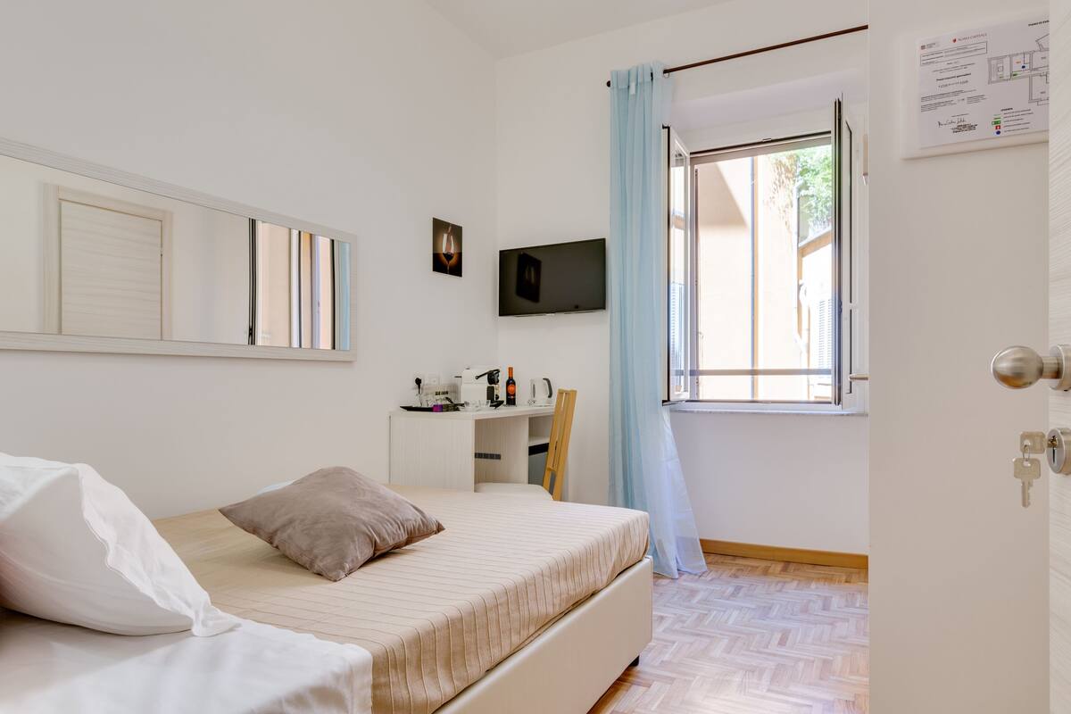 A bedroom with a bed, a desk, a TV, and a window with curtains.