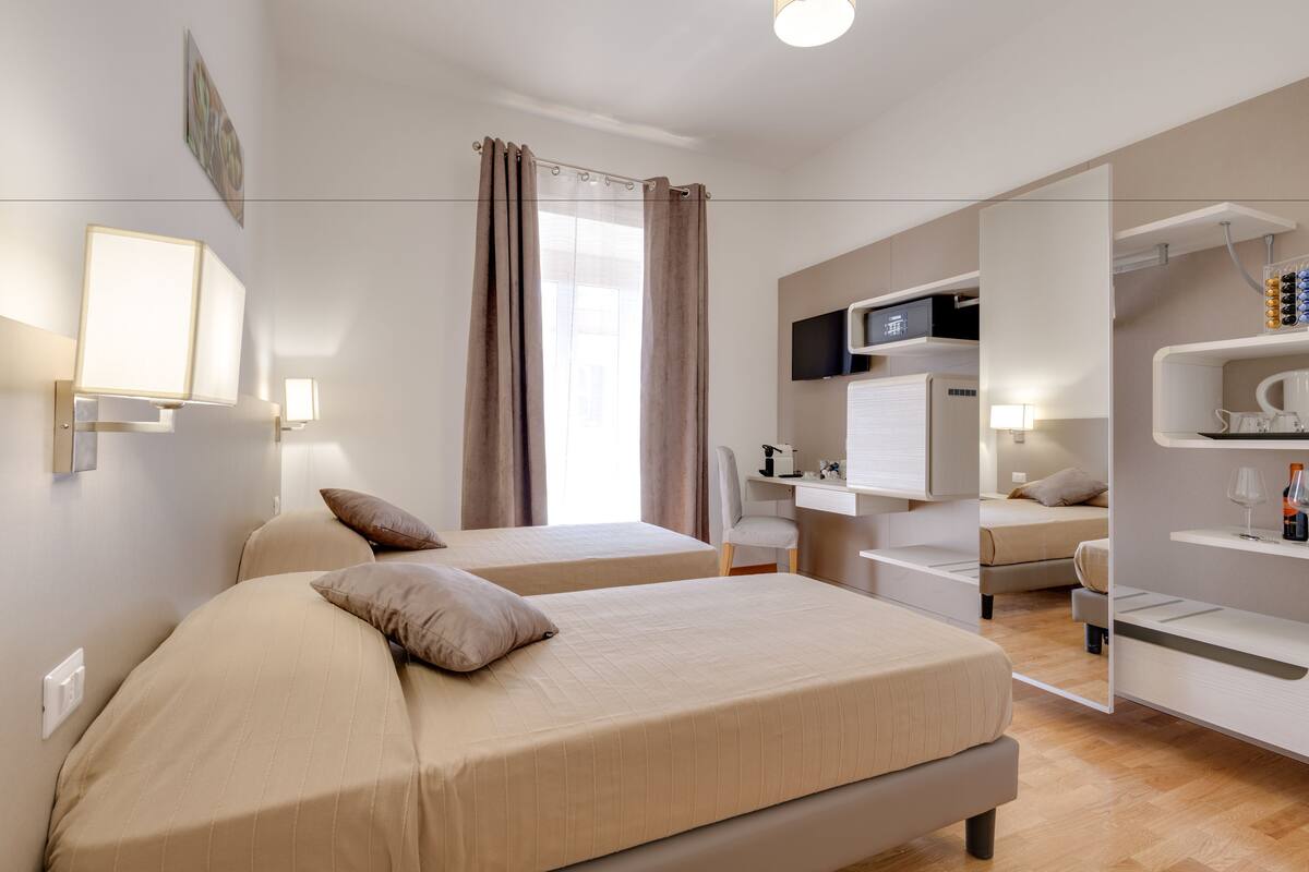 A modern hotel room with two beds, a desk, a chair, a TV, and a shelf with bottles.