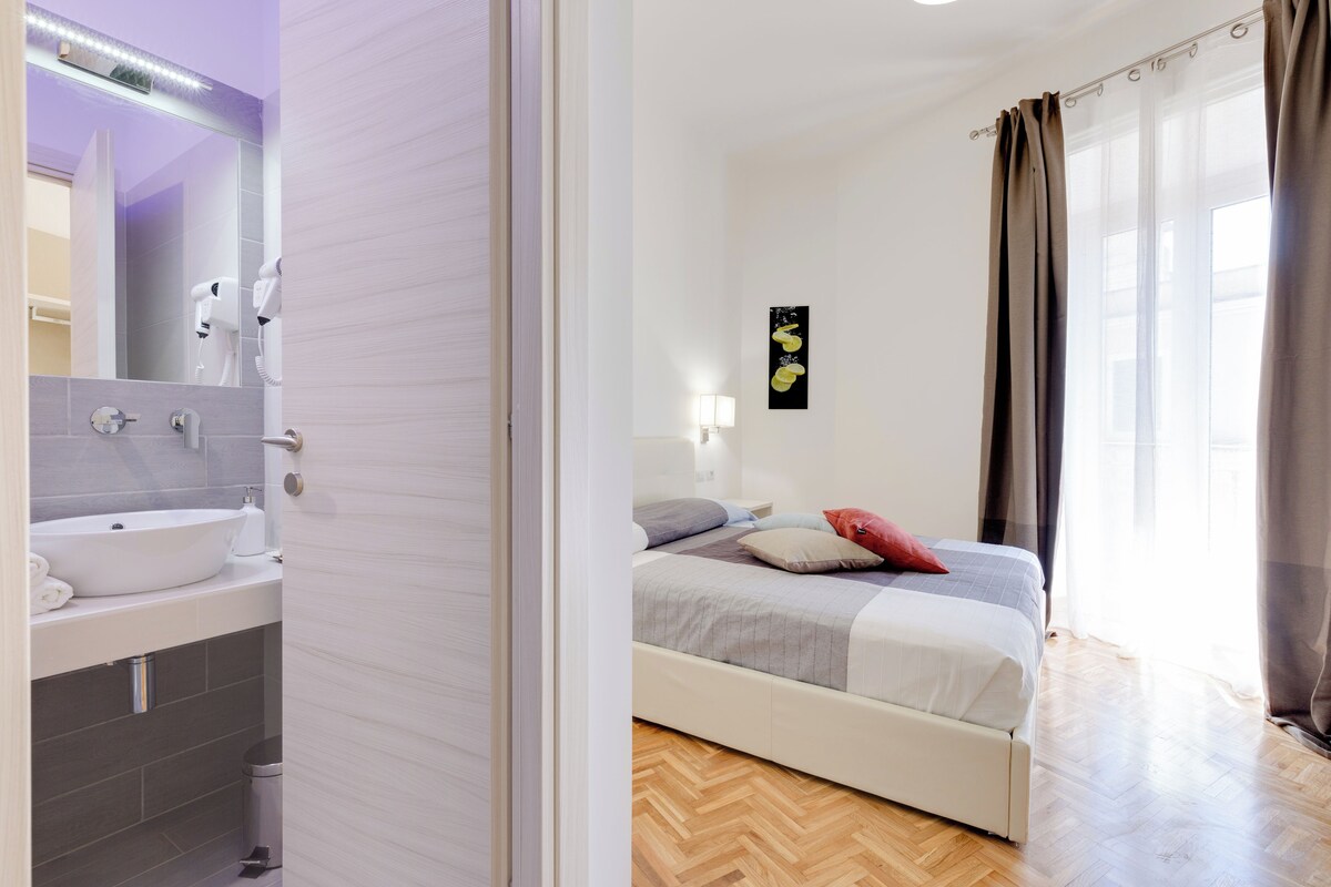 Een slaapkamer met een bed, nachtkastje, wandlamp en raam met gordijnen.