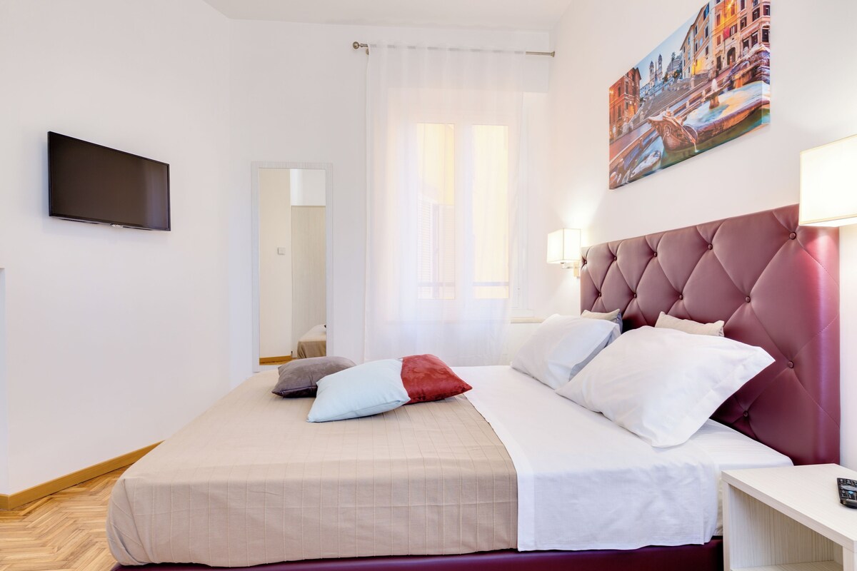 Habitación de hotel con cama, televisor montado en la pared, ventana con cortinas y un cuadro colgado.
