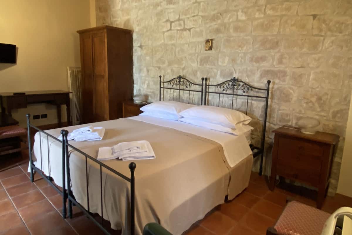 A room with a stone wall, a bed with white linens, a wooden wardrobe, and a small bedside table.