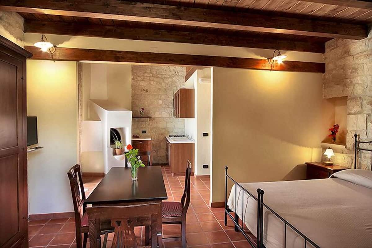 A cozy room with a bed, a dining table with chairs, a kitchen area, and a stone wall.