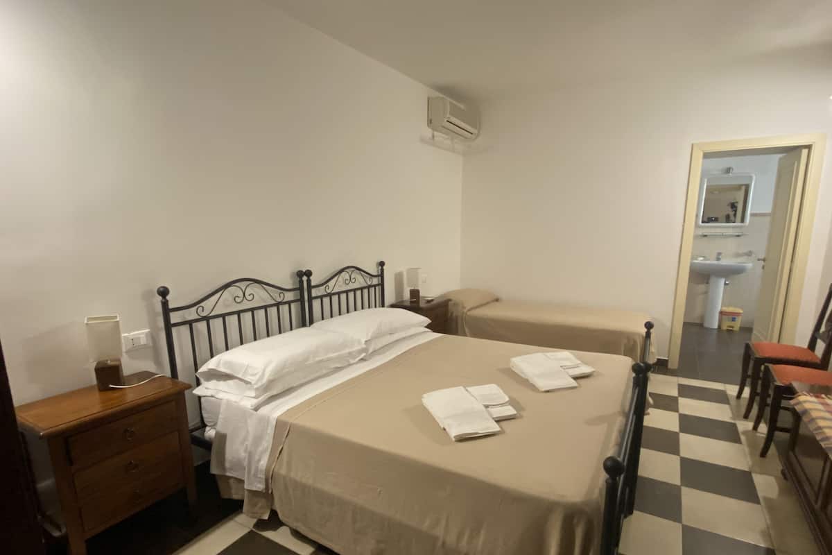 A hotel room with a checkered floor, a bed with a beige cover, a nightstand with a lamp, and a wooden dresser.
