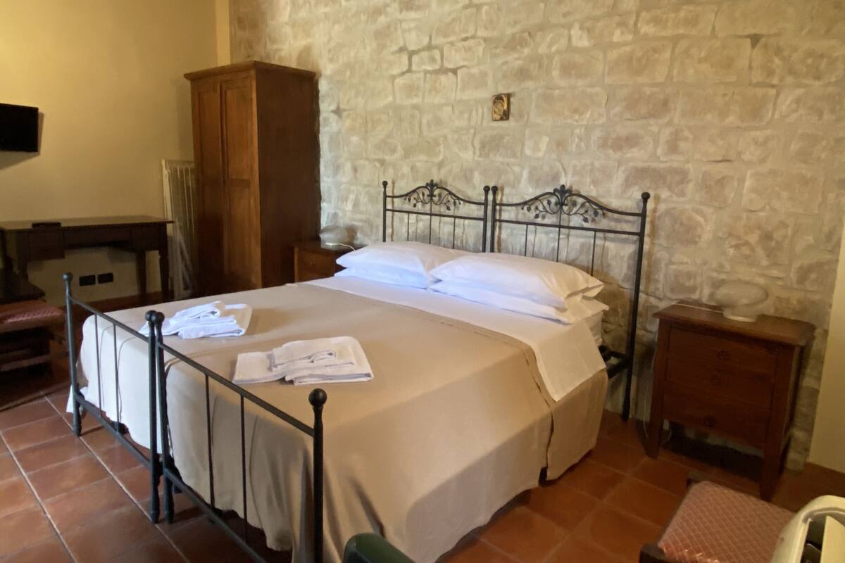 A bedroom with a stone wall, a metal bed frame, a wooden wardrobe, and a small bedside table.