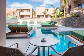 A pool area with lounge chairs and a table, surrounded by buildings with a modern design and palm trees. - Paralos Venus Suites - Adults Only (Hersonissos)