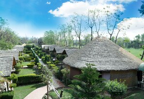 A series of thatched-roof huts arranged in a garden setting with a clear pathway leading through them. - The Corbett view resort (Ramnagar)