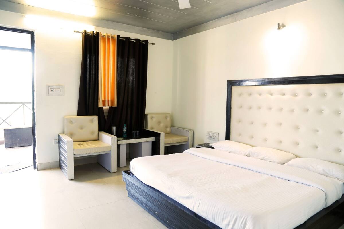 Quarto de hotel com uma cama grande, duas cadeiras, uma mesa pequena e uma janela com cortinas.