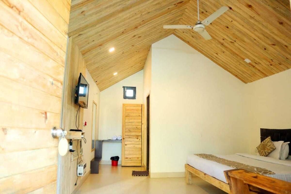 Um quarto com painéis de madeira, uma cama, um ventilador de teto, uma televisão e uma porta que dá para outro cômodo.
