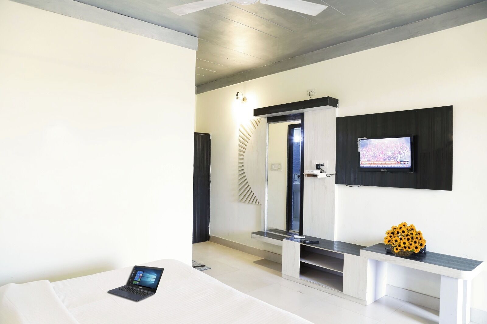 A modern hotel room with a bed, a laptop, a TV mounted on the wall, and a small bench with a vase of sunflowers.