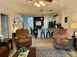 Una sala de estar con dos sillones marrones, una mesa de centro con una planta, y una cocina al fondo.