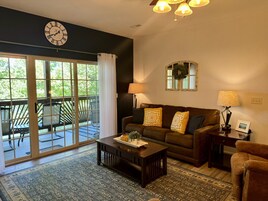 A living room with a sofa, coffee table, and lamps. There is a large window with curtains, a clock on the wall, and a view of the outdoors.