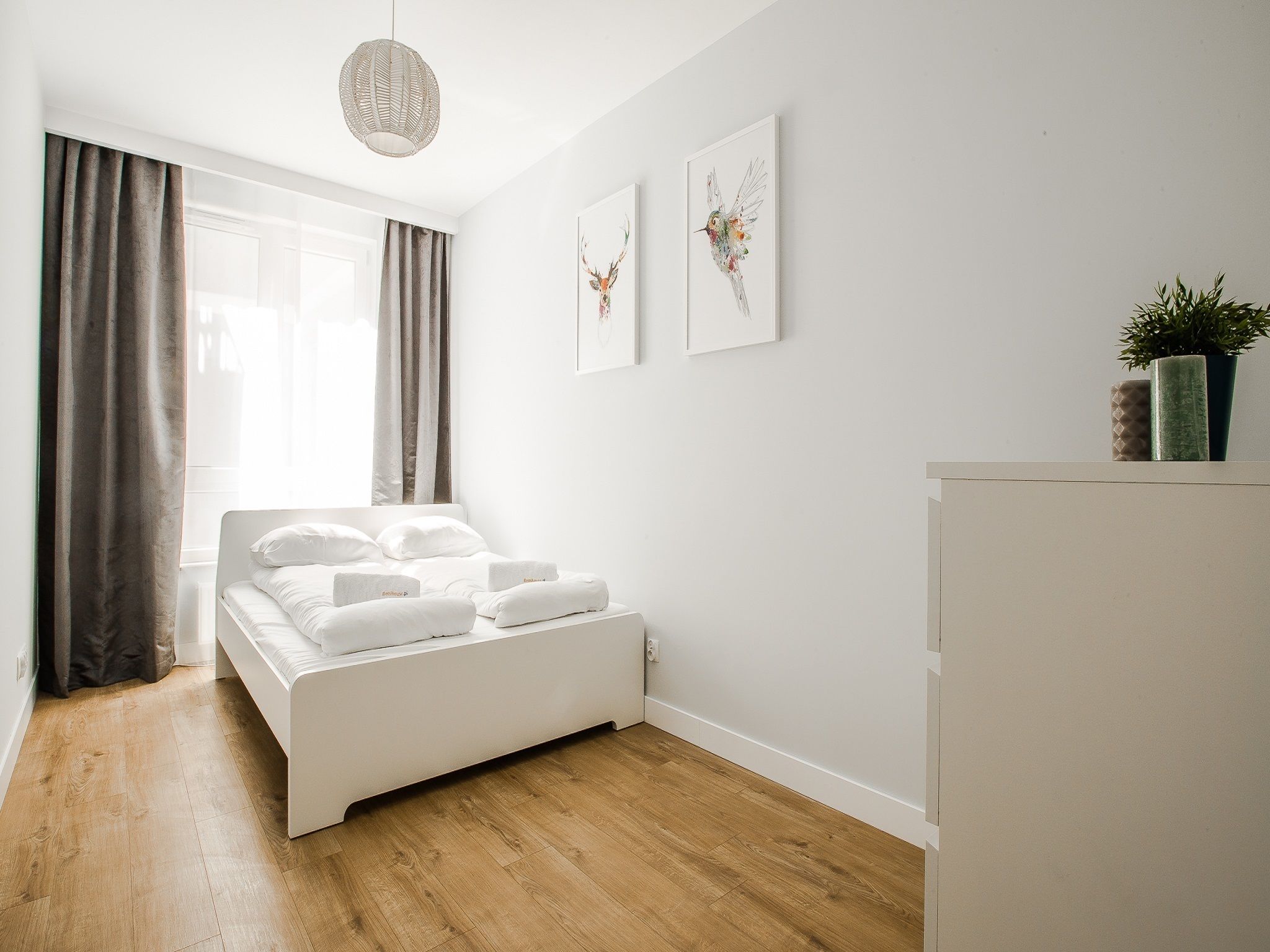 A modern bedroom with a bed, a bedside table, a hanging light fixture, and two wall-mounted artworks.