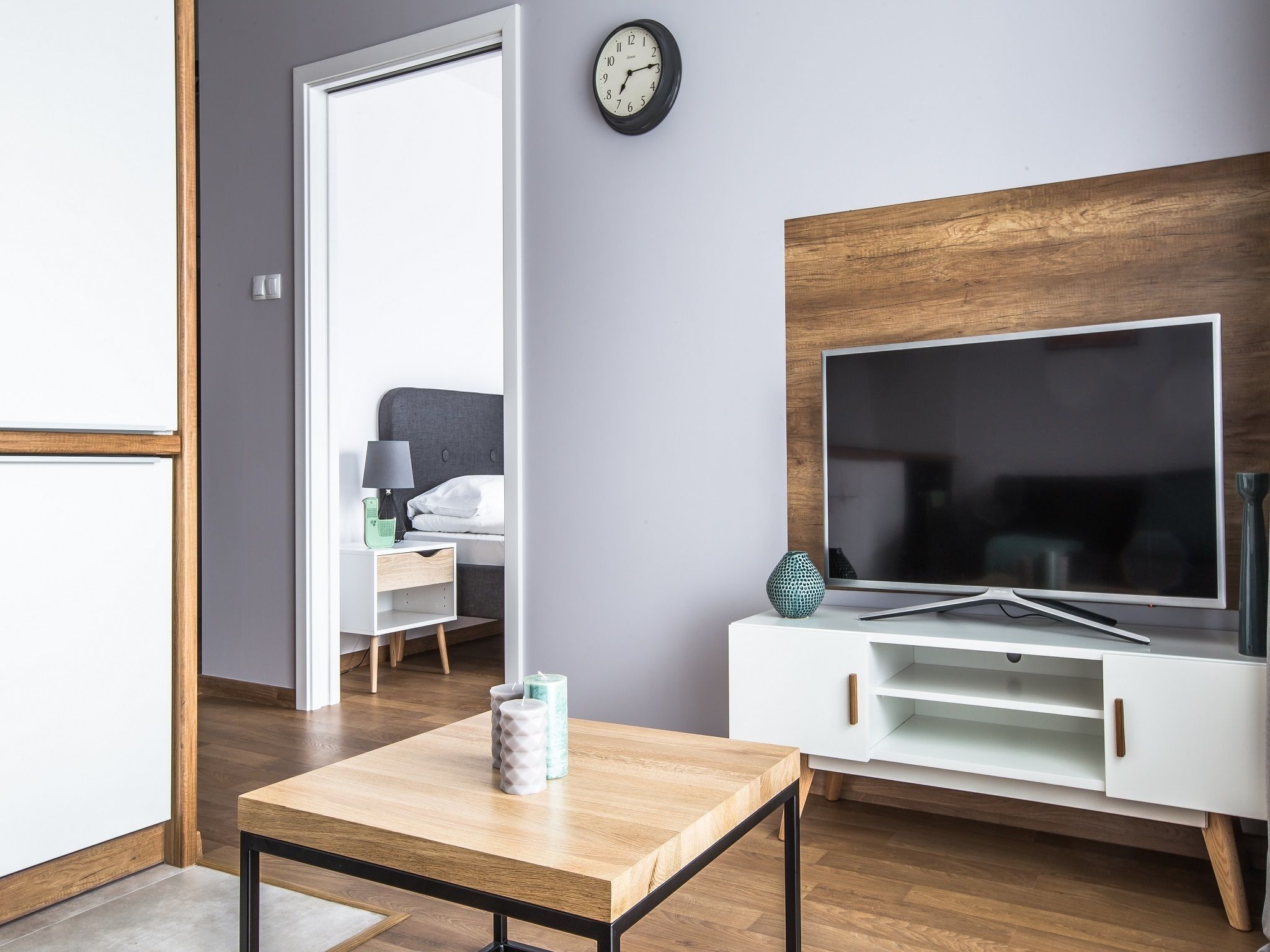 Ein modernes Wohnzimmer mit Holzboden, einem Fernseher auf einem weißen Schrank, einem kleinen Tisch mit Kerzen und einer Tür zu einem anderen Raum.