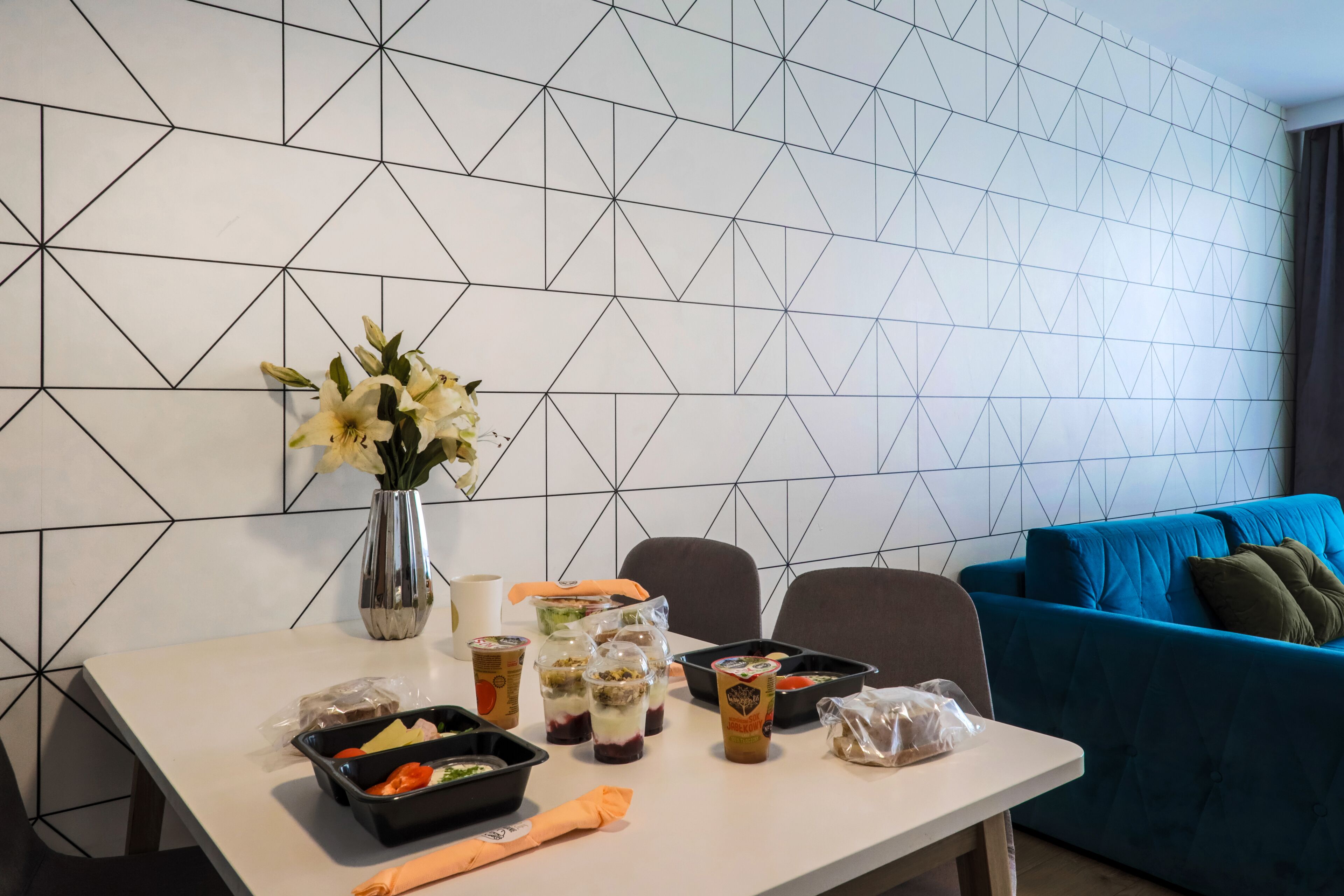 Een eettafel met maaltijdverpakkingen, een vaas met bloemen en een blauwe bank op de achtergrond.