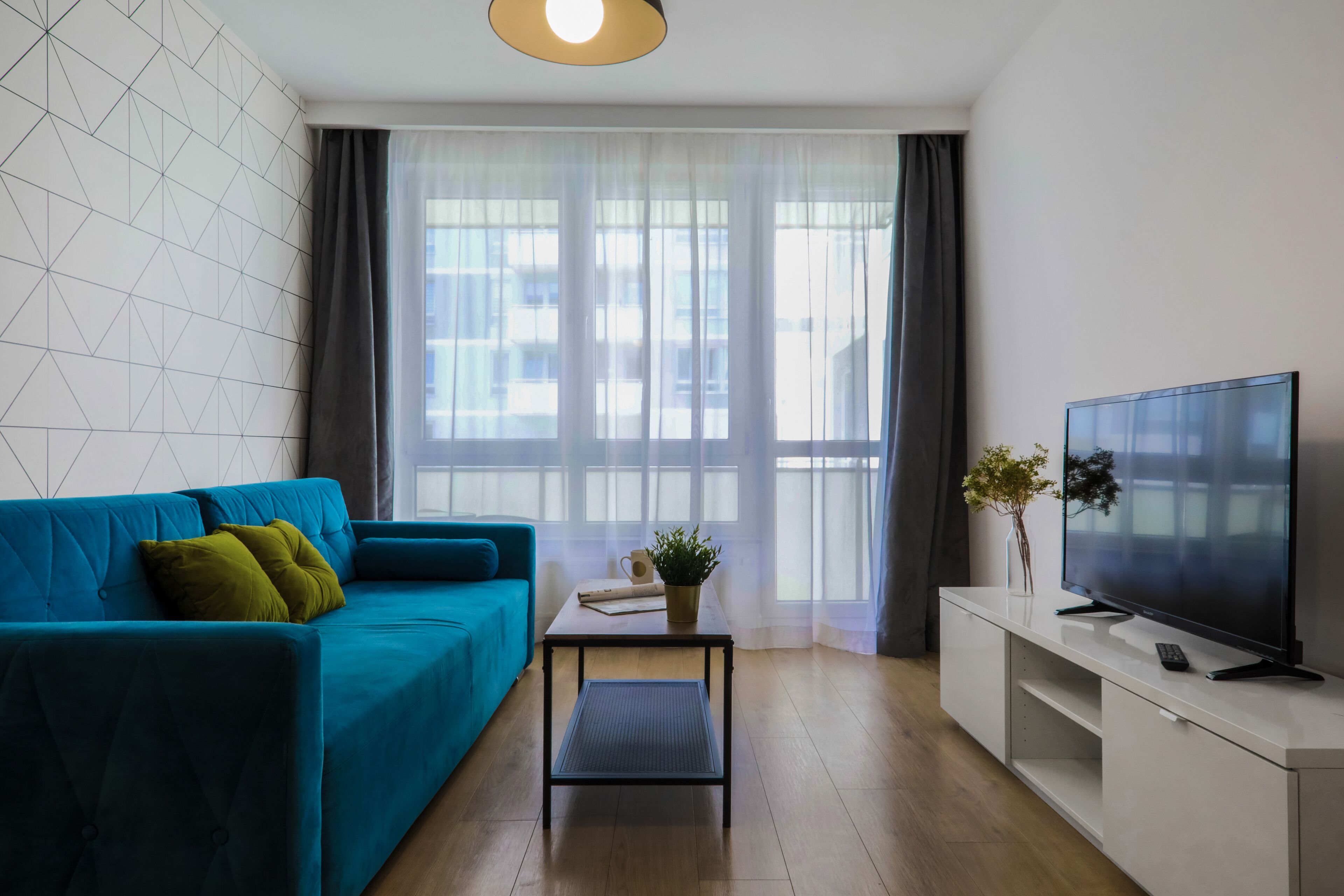A modern living room with a teal sofa, a small coffee table, a flat-screen TV, and a large window with sheer curtains.