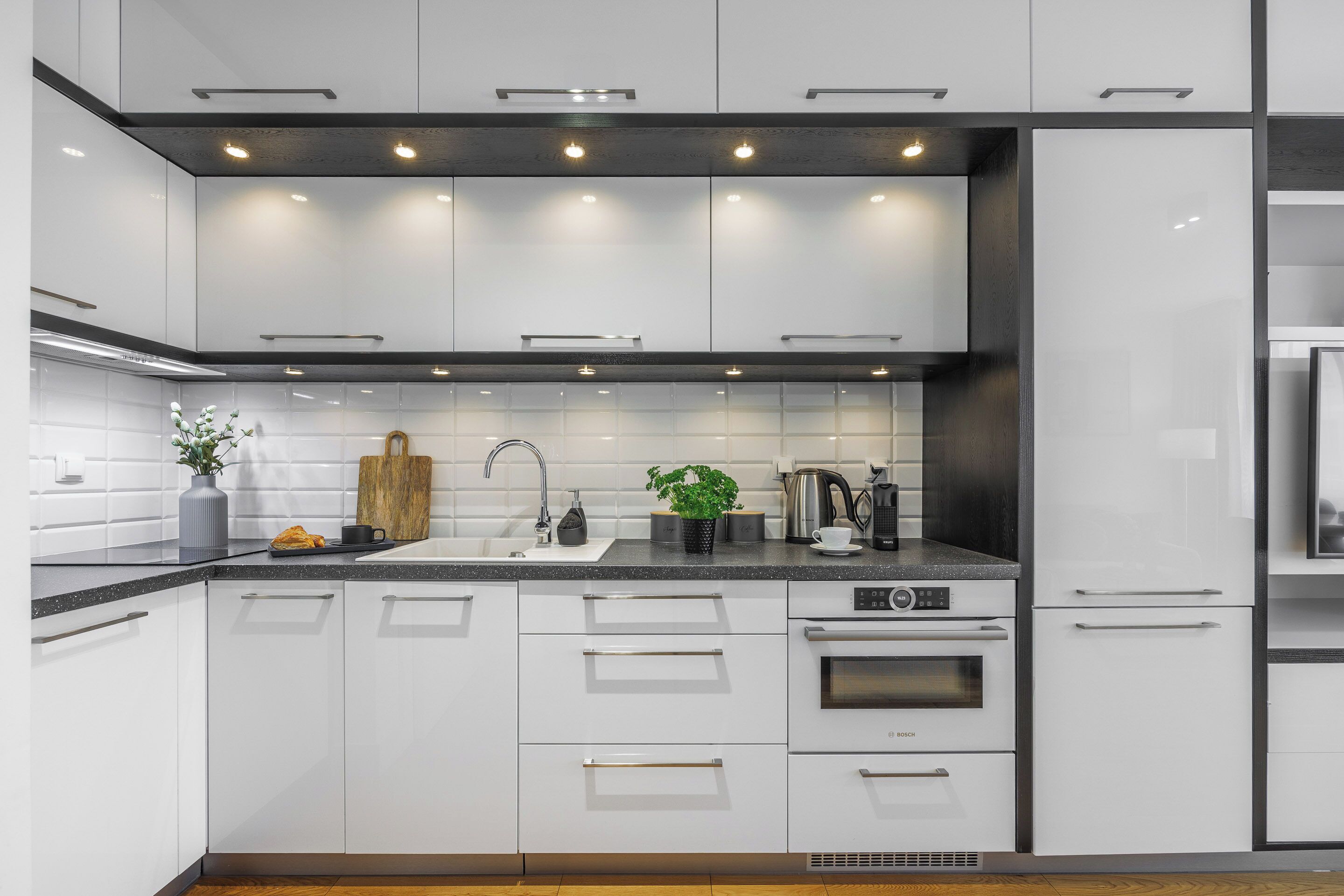 Eine moderne Küche mit weißen Schränken, einem Backofen von Bosch, einer Spüle und einer Arbeitsplatte mit Pflanze und Kaffeemaschine.