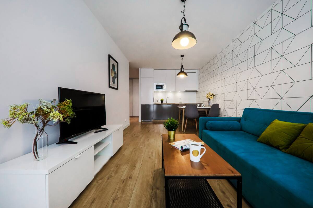 A modern living room with a flat-screen TV, a blue sofa, a coffee table, and a kitchen in the background.