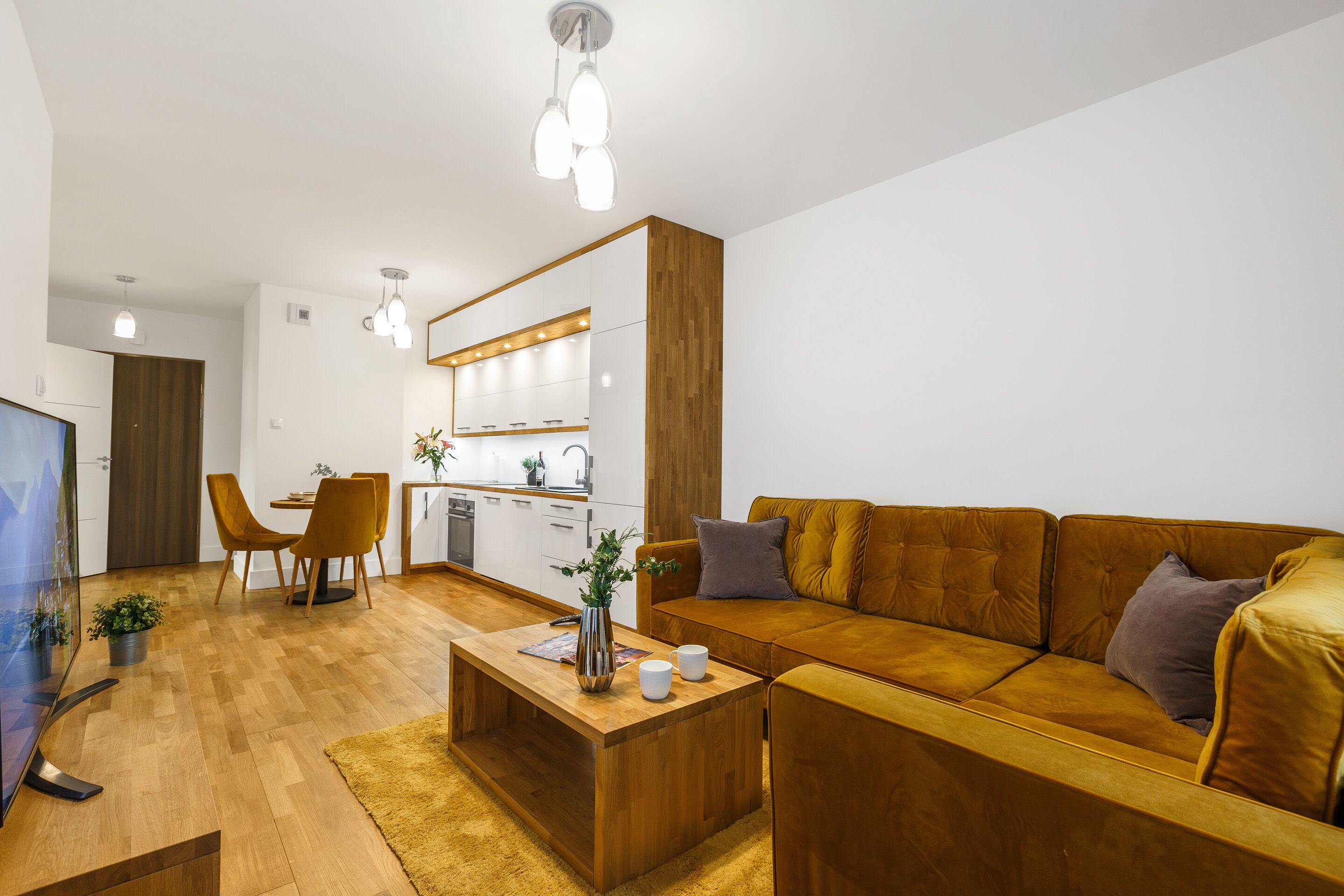 Una sala de estar moderna con un sofá de madera, una mesa de centro y un televisor. Hay una zona de cocina con armarios y una mesa de comedor con sillas.