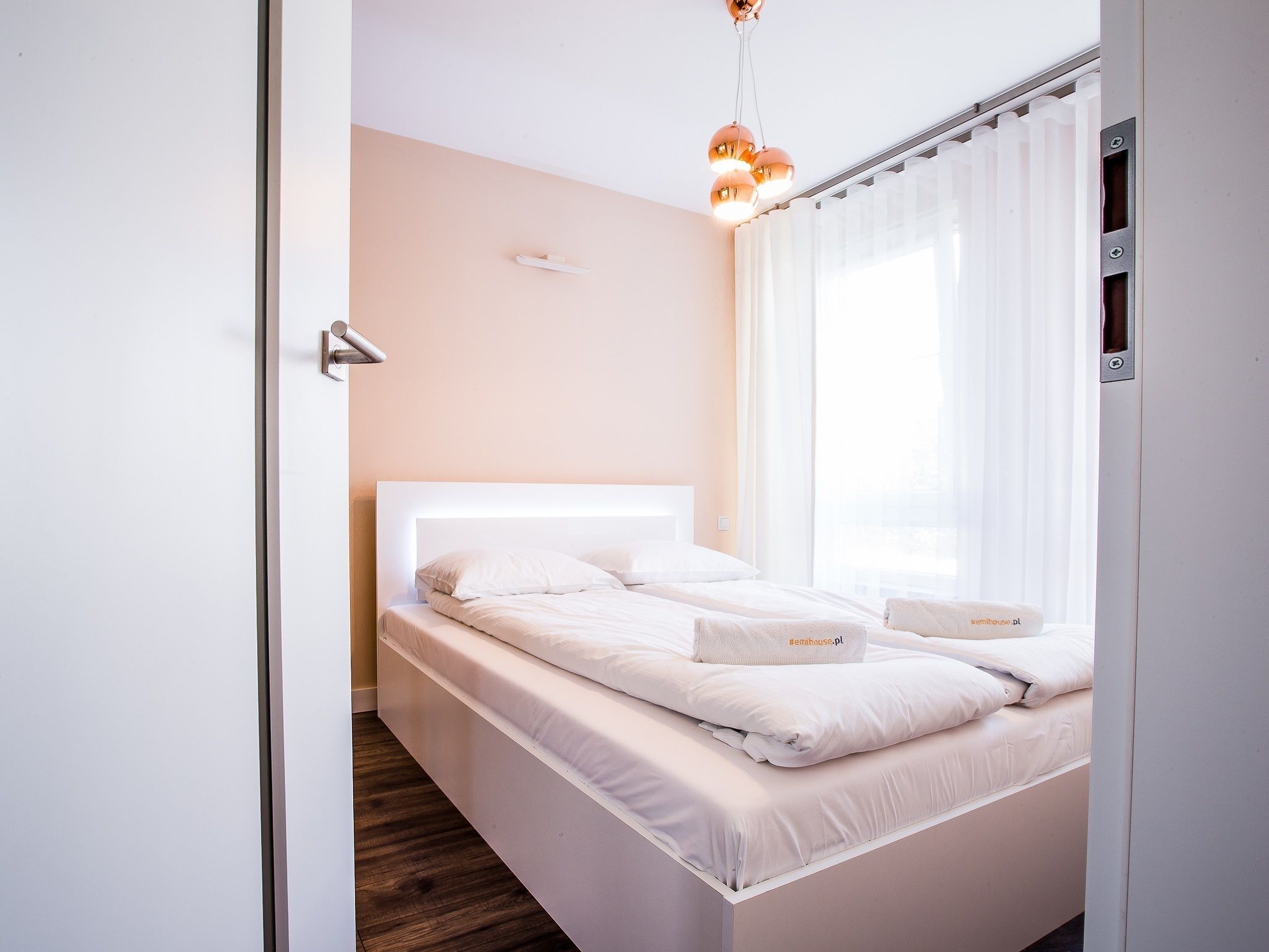 Un dormitorio moderno con una cama blanca, piso de madera, cabecera blanca y una lámpara en el techo.