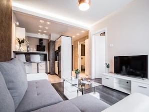 Una sala moderna con un sofá gris, una mesa de centro de vidrio, un televisor de pantalla plana y una zona de cocina con armarios blancos y un horno empotrado.
