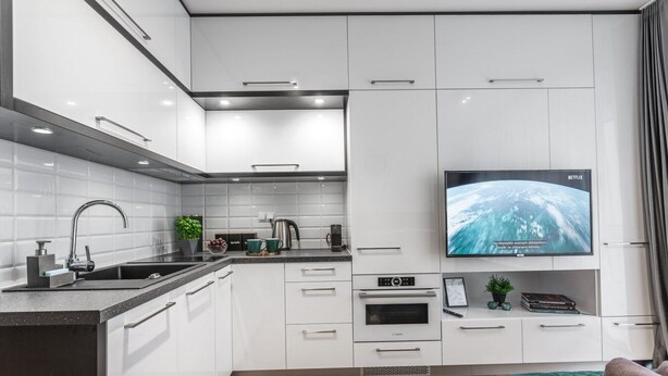 Eine moderne Küche mit weißen Schränken, einer schwarzen Arbeitsplatte und einem eingebauten Backofen. Ein Flachbildfernseher ist über dem Backofen angebracht und zeigt eine Naturansicht.