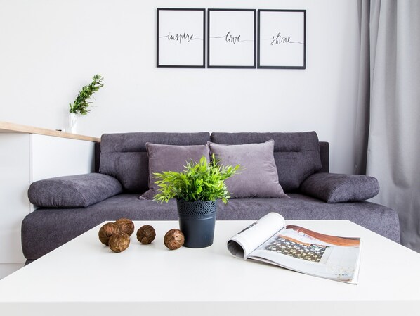 Una sala de estar moderna con un sofá gris, dos cojines, una planta en maceta y objetos decorativos sobre una mesa blanca.