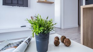 A modern living room with a large flat-screen TV mounted on the wall, wooden shelves with decorative items, a potted plant on a white table, and a magazine on the floor. - Emihouse CityVibe Apartments (Rzeszów)