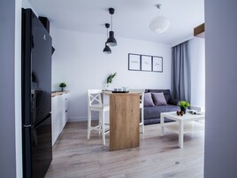 A modern kitchen with a black refrigerator, wooden dining table, and white chairs. There is a grey sofa and a white coffee table with a plant. The room has light wooden flooring and white walls.