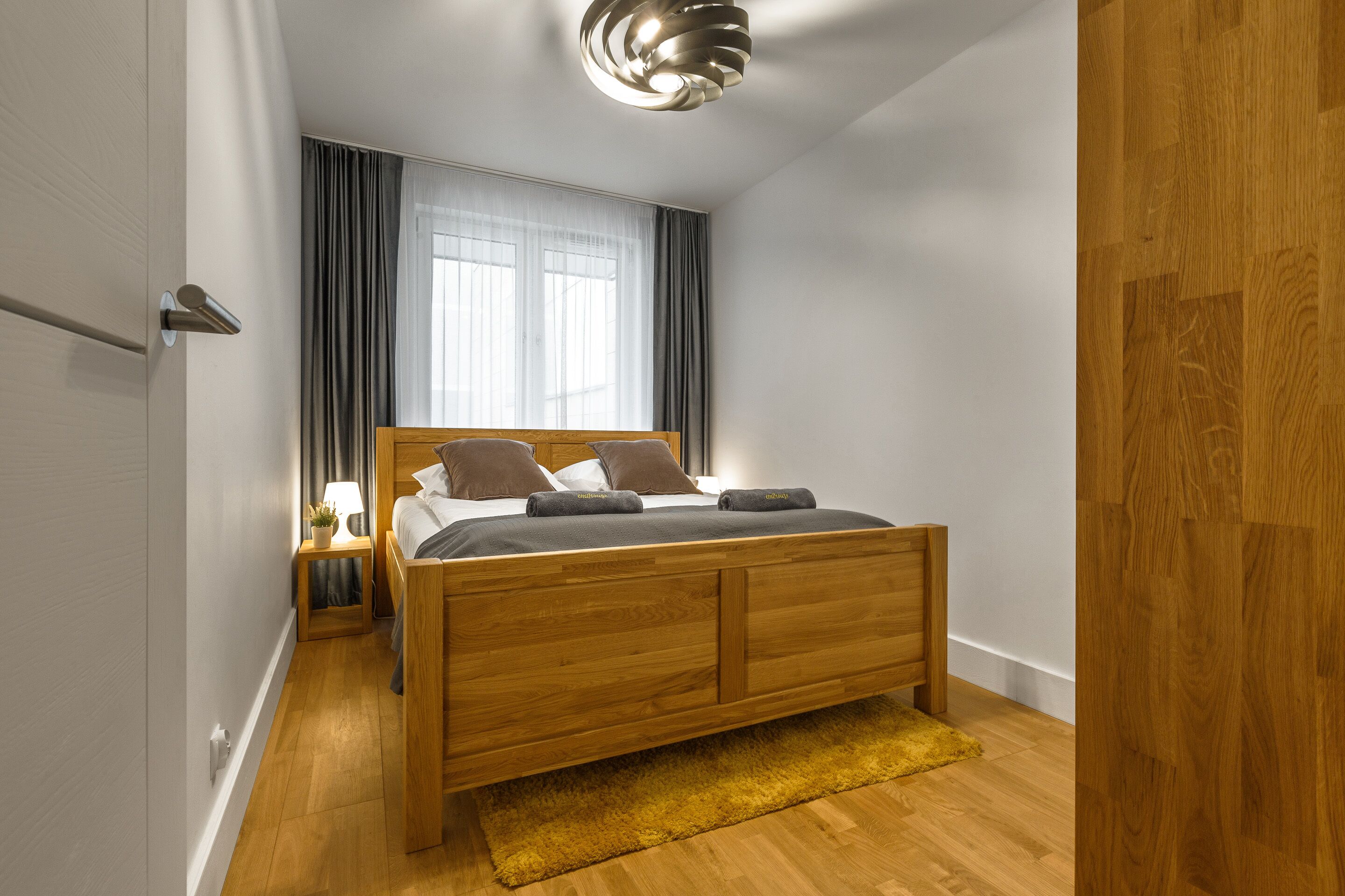 Un dormitorio moderno con una cama de madera, una mesita de noche, una lámpara y una ventana con cortinas.