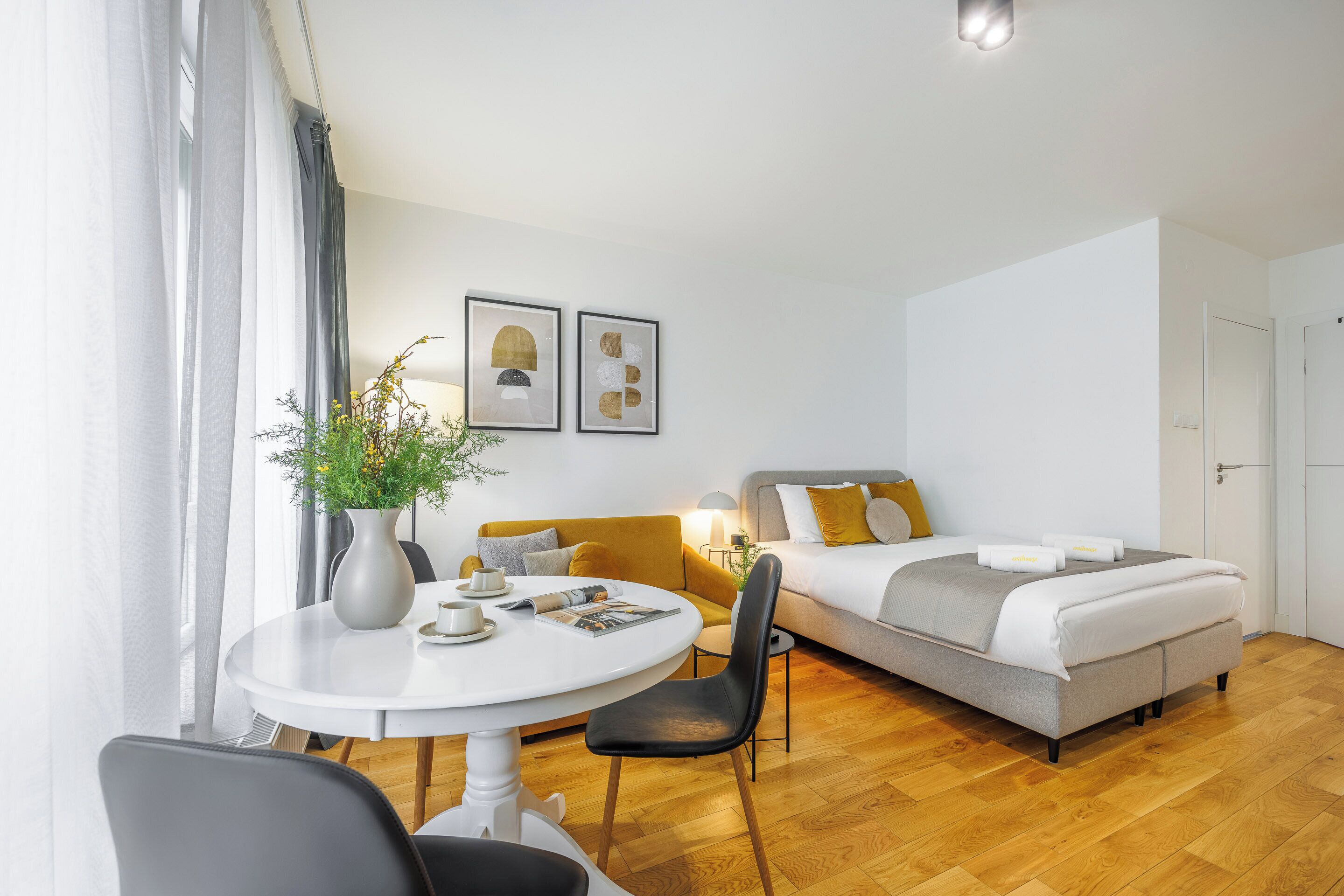 Habitación de hotel moderna con una mesa de comedor redonda, un sofá, una cama con decoración gris y blanca, y dos cuadros enmarcados en la pared.