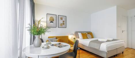 Habitación de hotel moderna con una mesa de comedor redonda, un sofá, una cama con decoración gris y blanca, y dos cuadros enmarcados en la pared.
