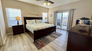 A bedroom with a bed, a ceiling fan, a dresser, a nightstand, a lamp, a rug, a window with blinds, a clock, and a view of the outdoors through a sliding glass door. - Walk-in Condo A-4 with Overlooking Deck by RedAwning (Hollister)
