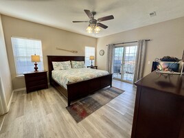 Un dormitorio con cama, ventilador de techo, cómoda, mesita de noche, lámpara, alfombra, ventana con persianas, reloj y vista al exterior a través de una puerta corrediza de vidrio.