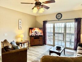 Una sala de estar con chimenea, ventilador de techo, televisor y vistas a través de un ventanal.