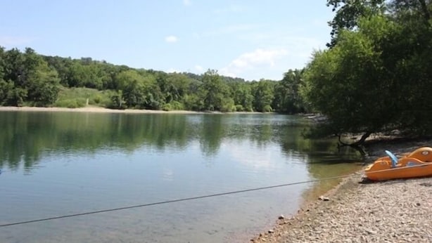 A calm lake with a boat on the shore and a forested hill in the background. - Walk-in Condo A-4 with Overlooking Deck by RedAwning (Hollister)
