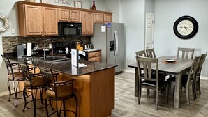 A modern kitchen with wooden cabinets, a central island with bar stools, a dining table with chairs, and a refrigerator.