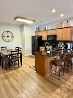 Una cocina moderna con armarios de madera, un refrigerador negro, un microondas y un comedor con una mesa y sillas de madera.