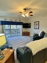 A living room with a ceiling fan, a TV, a couch, a chair, a lamp, and a view of the outdoors.