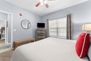 Une chambre à coucher comprenant un lit, une commode, un téléviseur, un miroir et une fenêtre avec des stores.