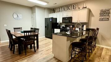 A modern kitchen with white cabinets, a black refrigerator, and a microwave. There is a wooden dining table with chairs and a clock on the wall.