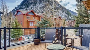 Une terrasse en bois, meublée de meubles en osier, avec une petite table et une vue sur des montagnes et des bâtiments enneigés.
