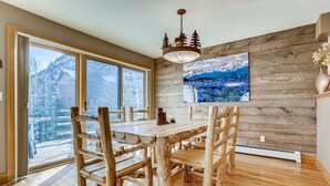 Une table à manger en bois, accompagnée de six chaises, de grandes fenêtres offrant une vue sur un paysage enneigé, un mur en bois orné d’un tableau représentant une montagne et un luminaire suspendu.