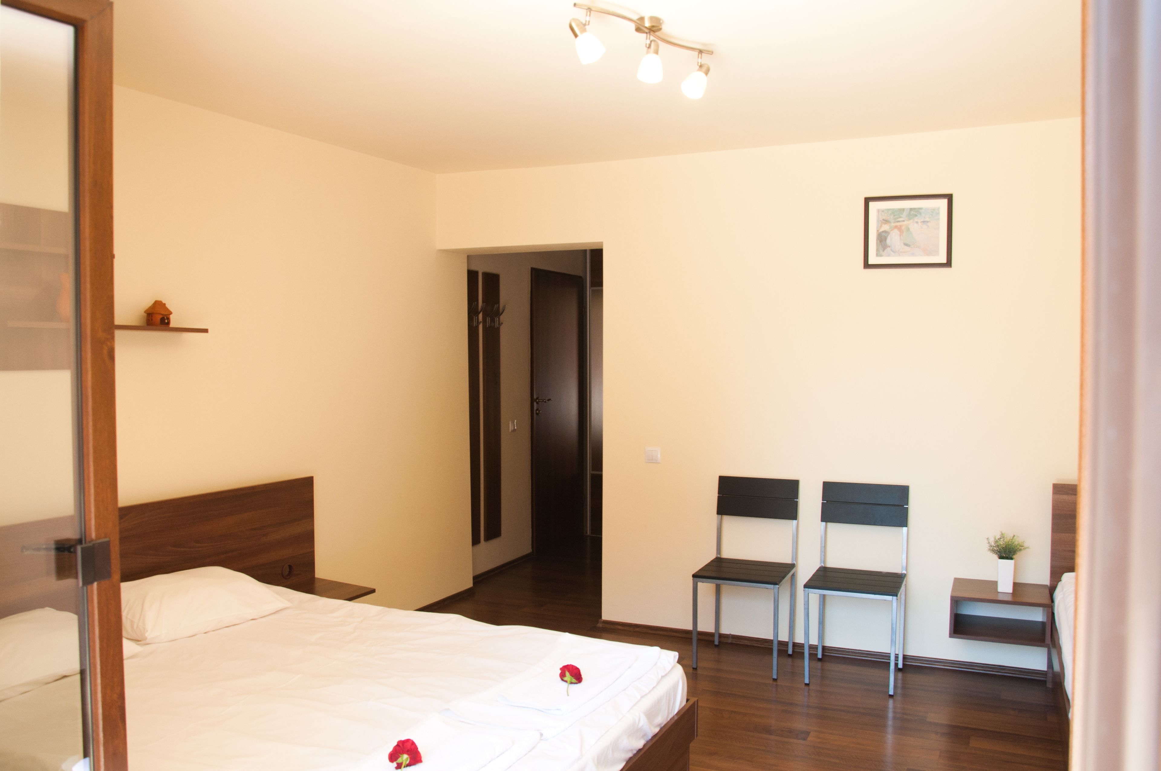 Una habitación de hotel con una cama de madera, una mesita pequeña, dos sillas, una puerta y un cuadro enmarcado en la pared.
