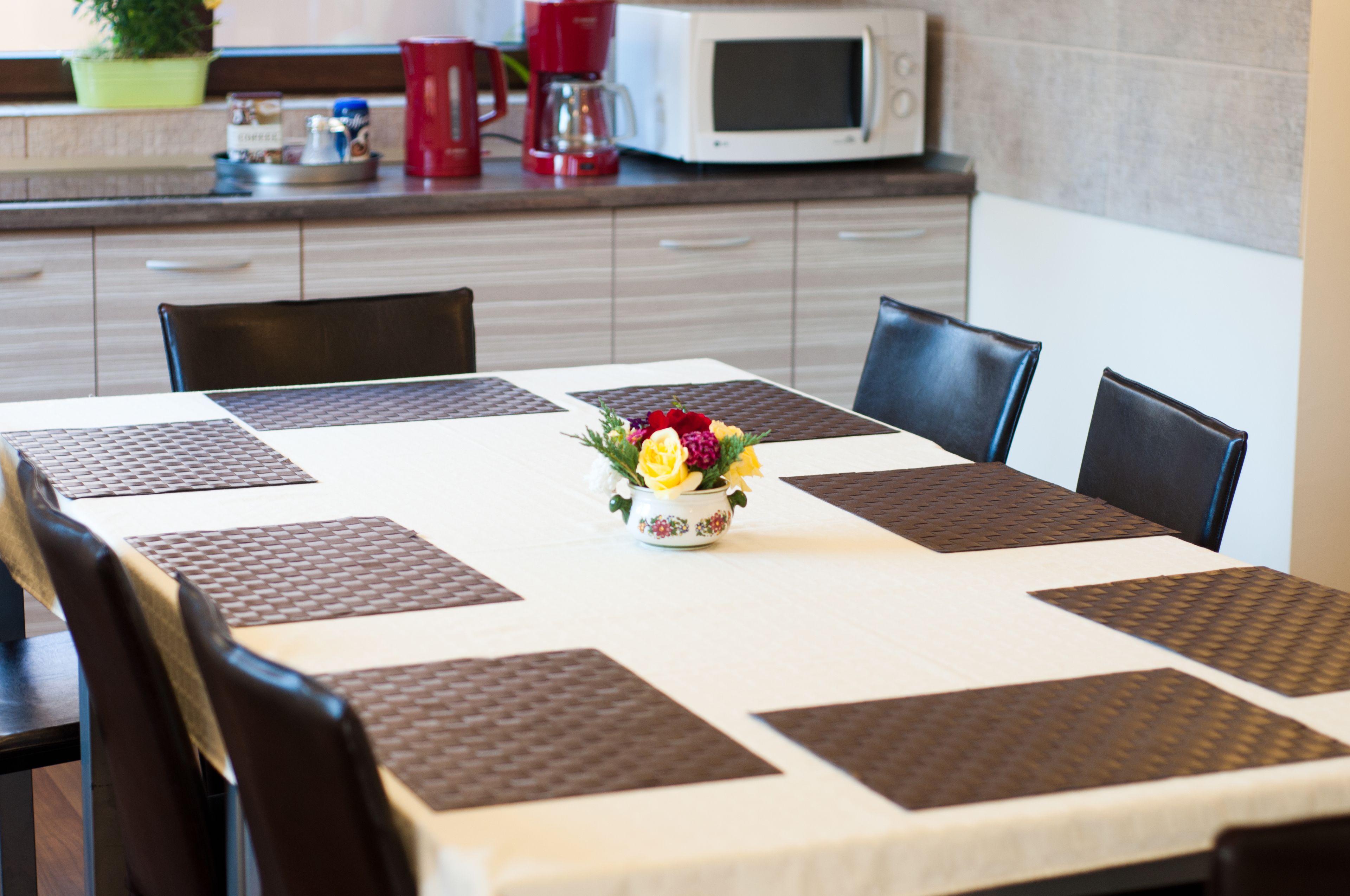 Ein Esstisch mit weißer Tischdecke und braunen Platzsets, umgeben von schwarzen Stühlen. In der Mitte steht eine florale Tischdekoration.