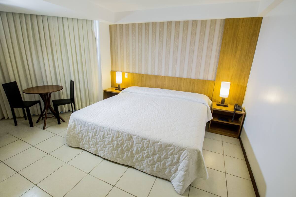 Habitación de hotel con una cama grande, dos mesitas de noche con lámparas, una pequeña mesa redonda con dos sillas y una ventana con persianas.