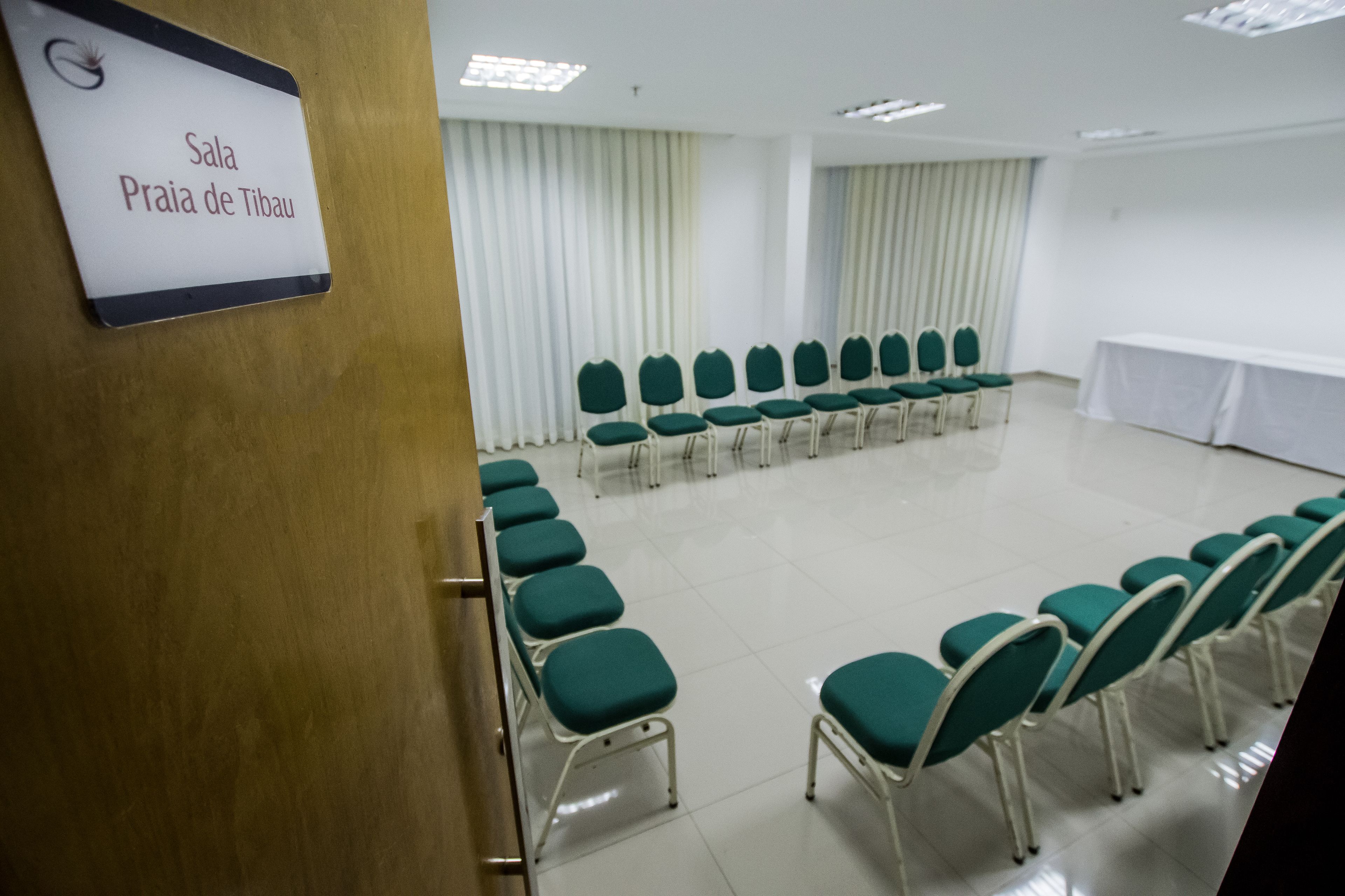 Un salón de conferencias con filas de sillas verdes y una mesa blanca, un cartel que dice 'Sala Praia de Tibau'.